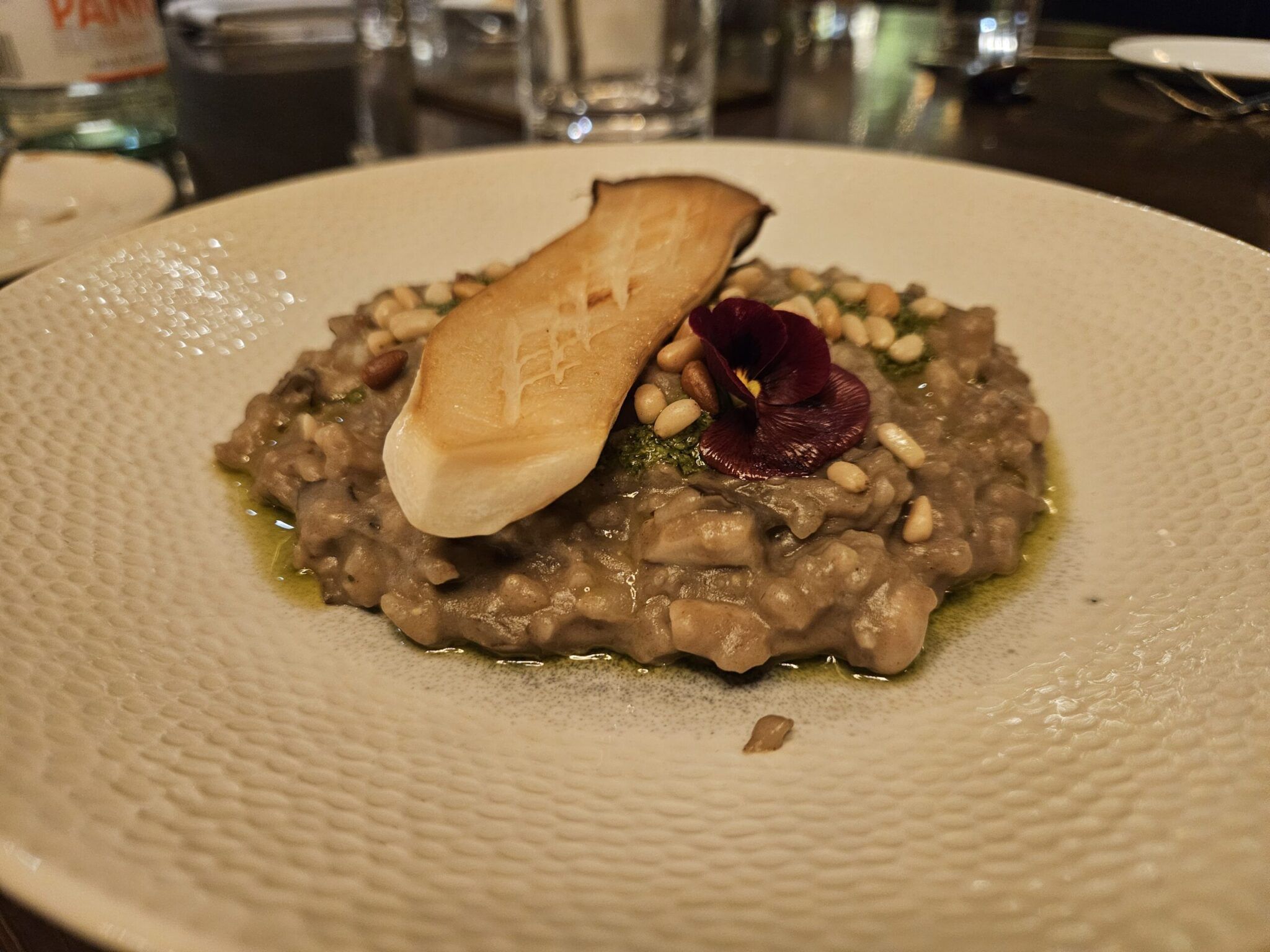 Mushroom risotto topped with seared king oyster mushroom, pine nuts, herb oil and a purple pansy on a textured plate