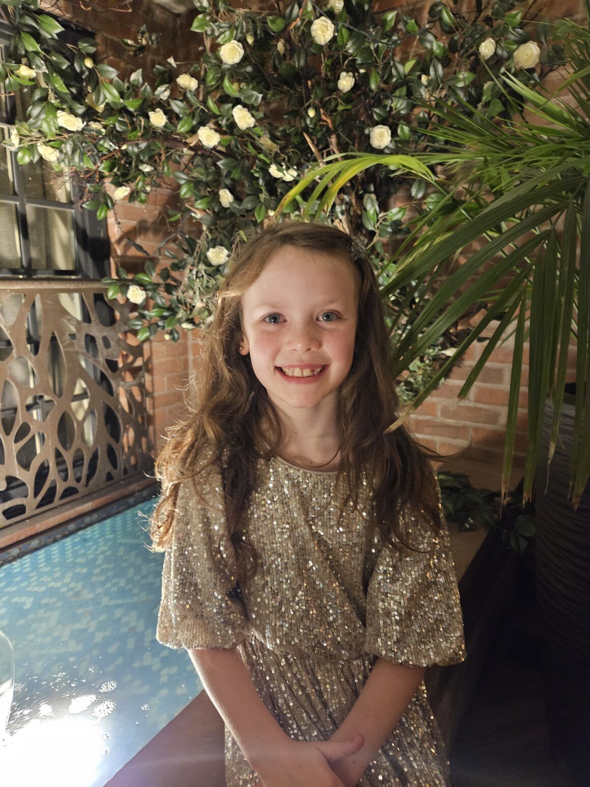 Smiling girl in a gold sequin dress beside an indoor pool, with white flowers and palm leaves against a brick wall.