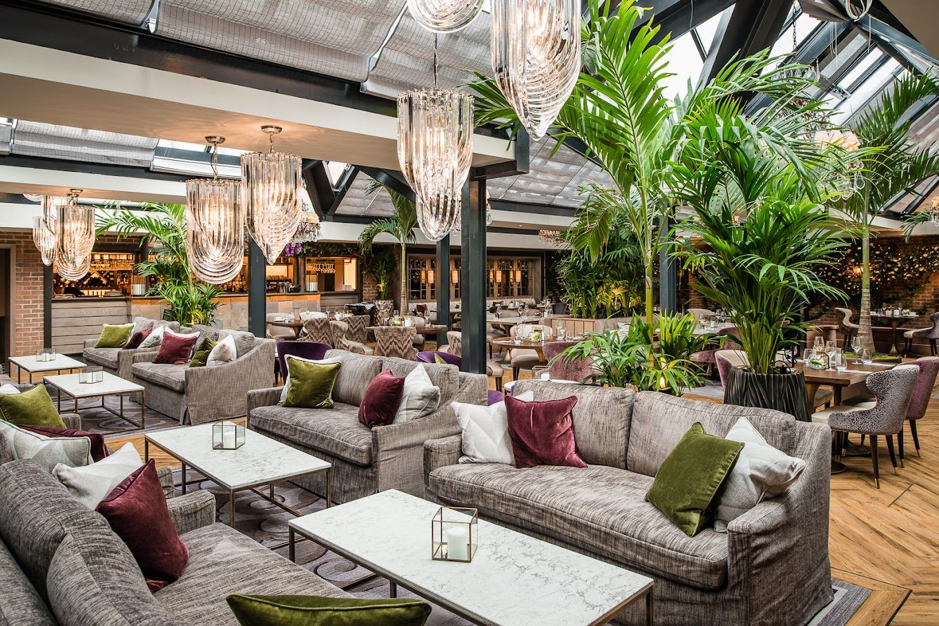 Glass-roofed hotel lounge with grey sofas, velvet cushions, marble tables, crystal chandeliers and tall palms.