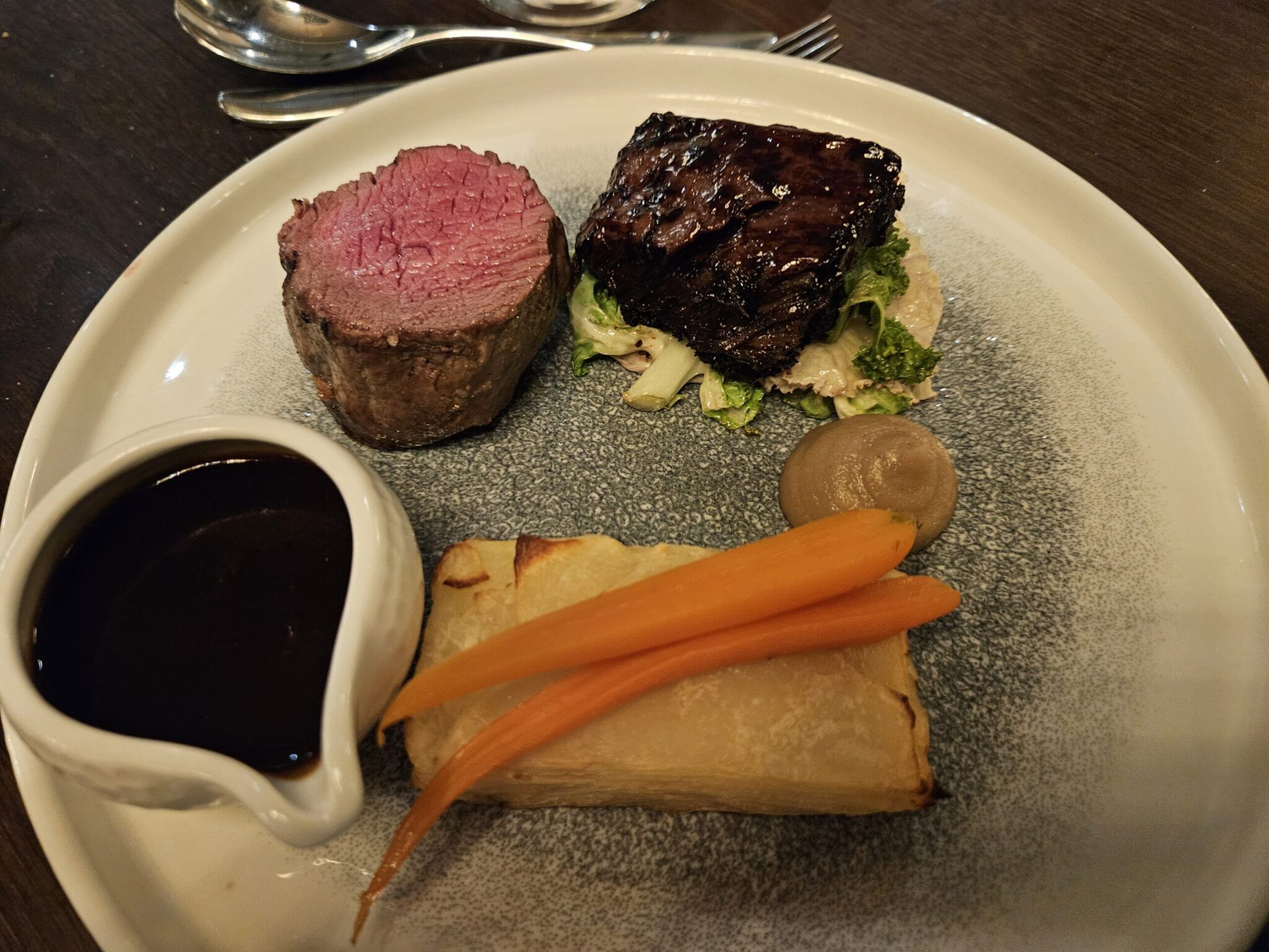 Plated beef fillet and glazed short rib with potato terrine, baby carrots, purée and a jug of jus on a speckled plate.