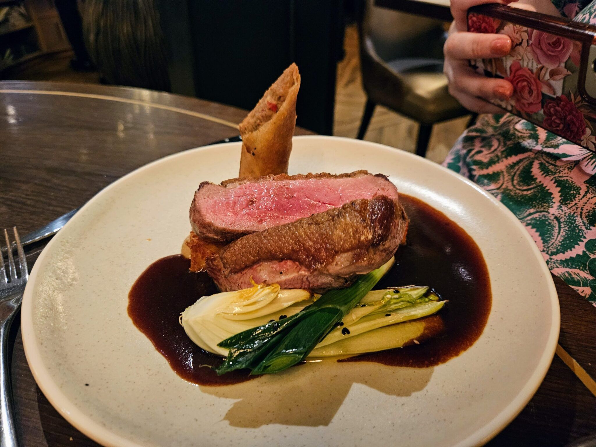 Sliced pink duck breast with a crispy spring roll, pak choi and dark jus on a white plate; a diner photographs it.