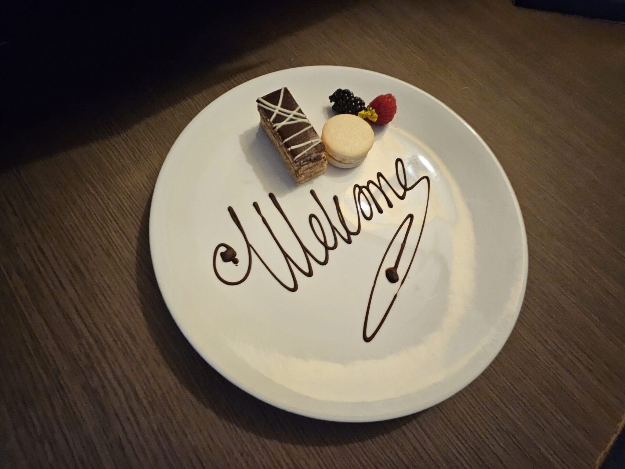 White plate with "Welcome" written in chocolate, a layered chocolate slice, macaron, raspberry and blackberry.