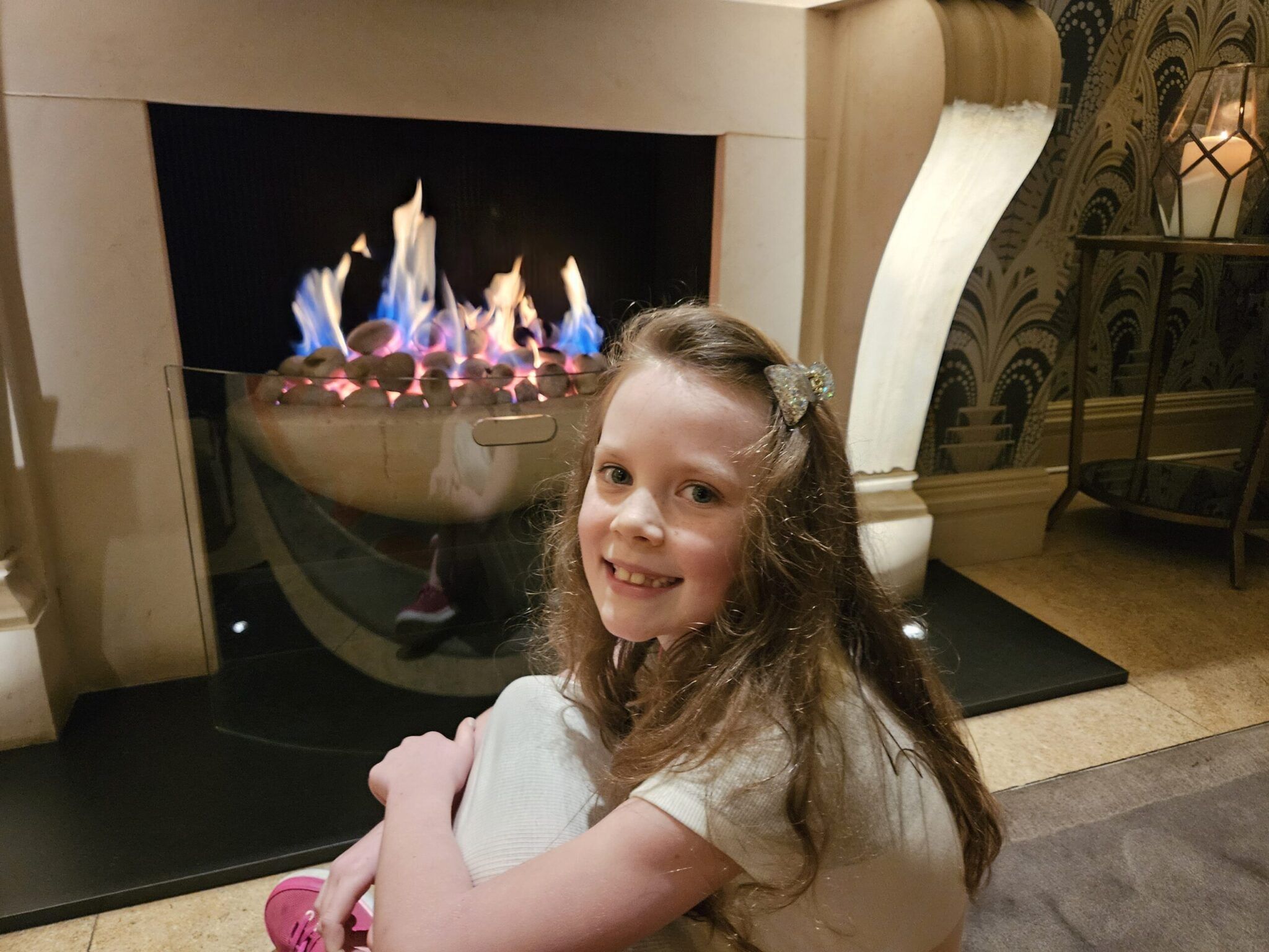 Smiling girl with long hair and sparkly bow sits by a stone fireplace with blue flames in a hotel lounge.