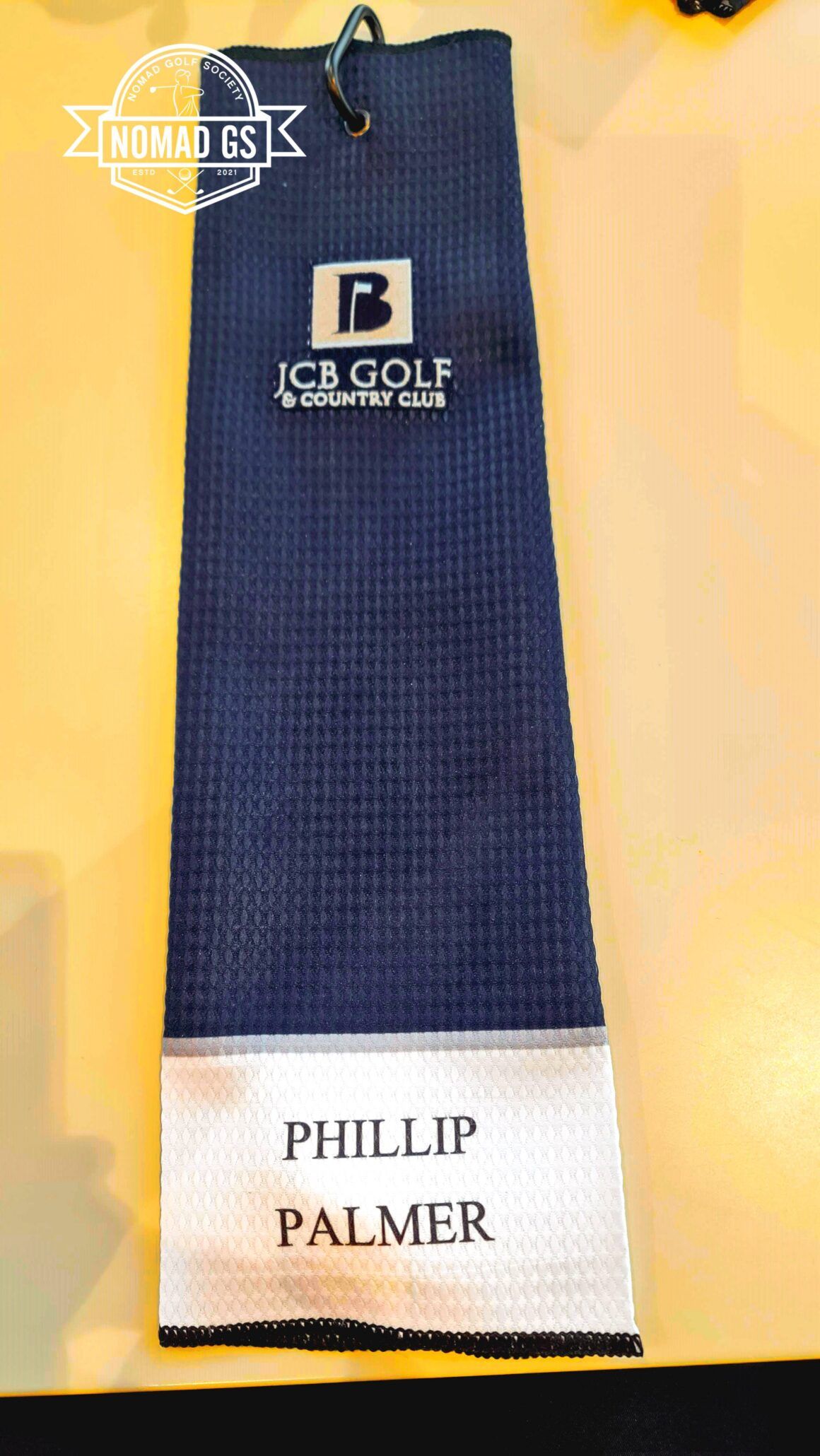Navy JCB Golf & Country Club towel with carabiner clip, personalised "Phillip Palmer" on white panel, on a yellow table.
