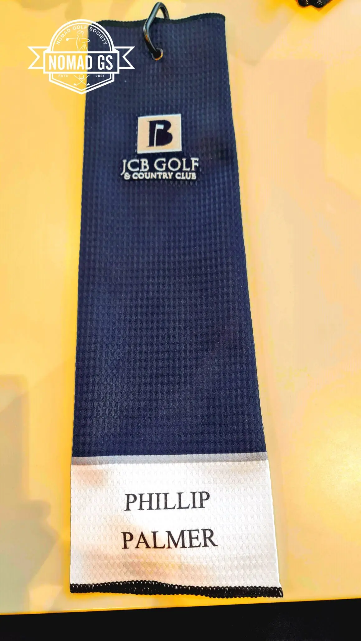 Navy JCB Golf & Country Club towel with carabiner clip, personalised "Phillip Palmer" on white panel, on a yellow table.