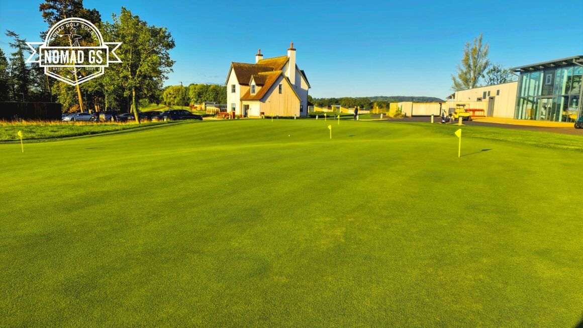 Large, immaculate putting green with yellow flags in front of JCB clubhouse buildings under a clear blue sky.