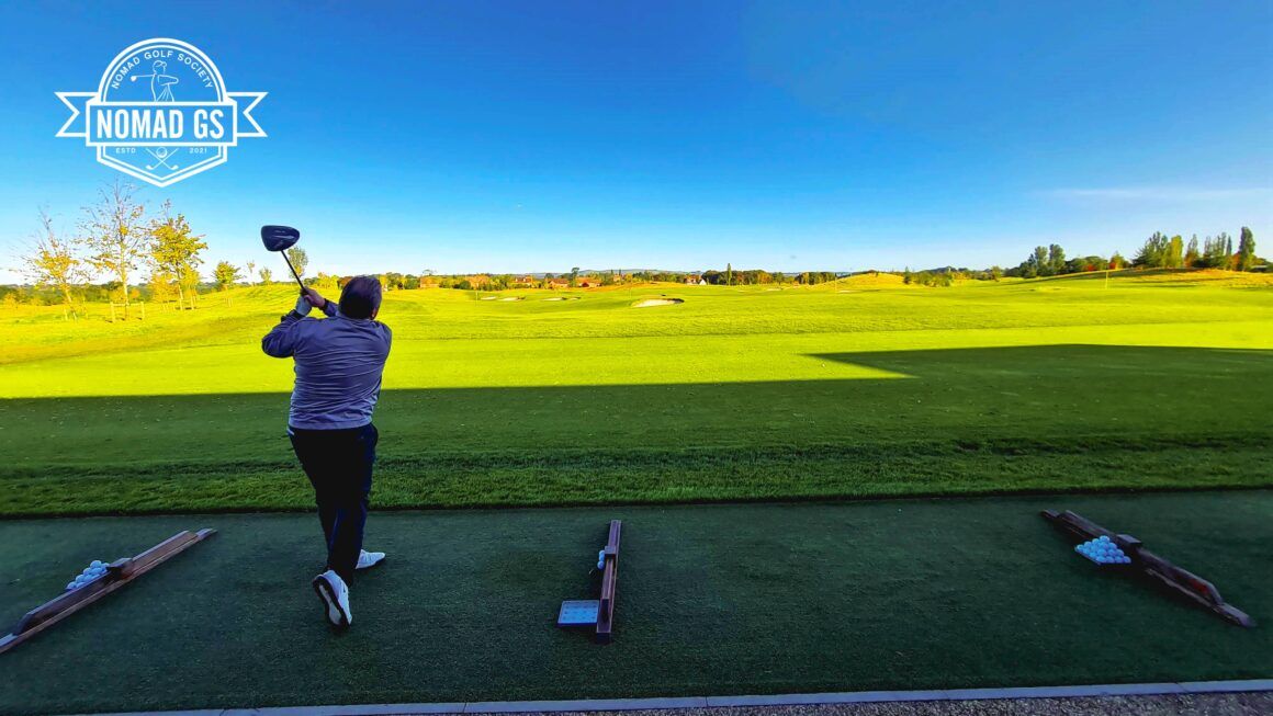 Golfer finishing a driver swing on JCB's grass driving range under clear blue sky, ball trays lined up beside him.